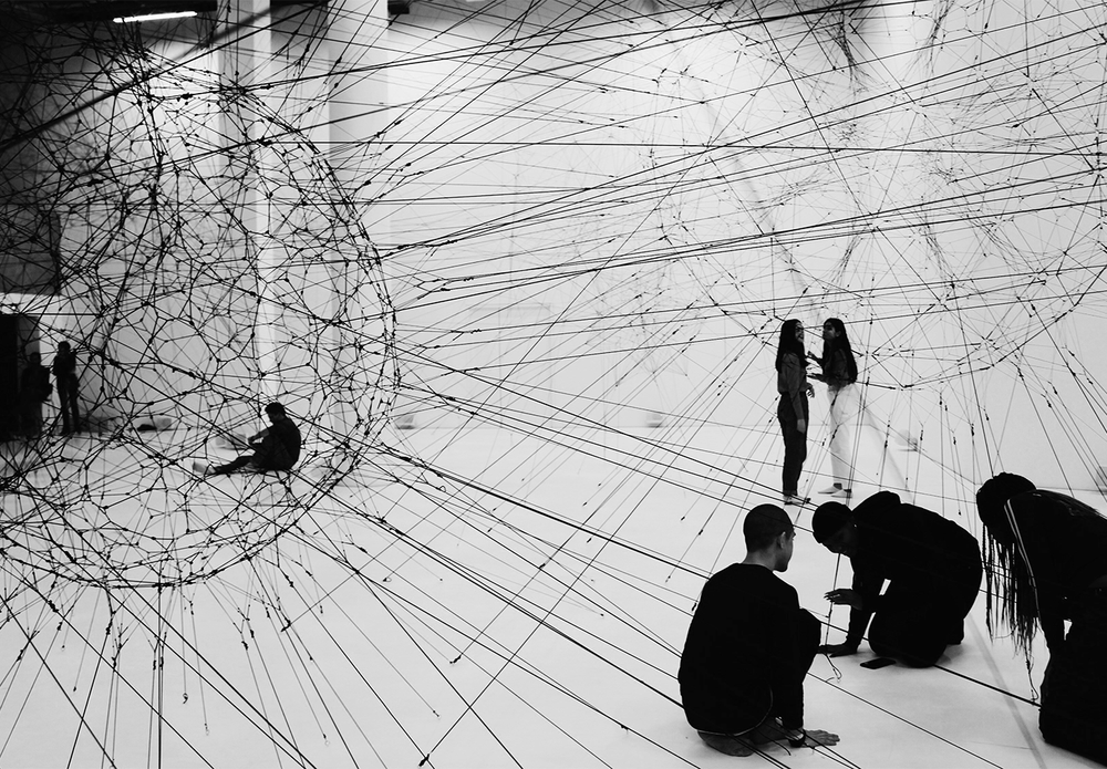 An art installation comprising of threads and strings and people on a white floor working on it,